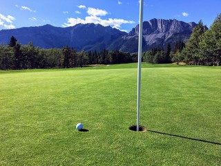 Obraz premium A view of a golf ball on a green close to the hole with the mountains and forest in the background on a beautiful summer day.