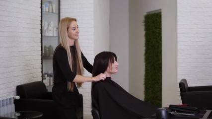 Female hairdresser has just finished working on her female client with long, straight brown hair. Woman is smiling and glad about her new hair style. Hairdresser removing protective cape. Side view