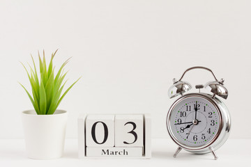 March 3 on a wooden calendar next to an alarm clock and a flower on a white background. Spring in the yard