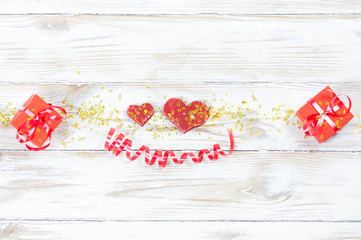 two red wooden hearts and two red gift boxes with bows in the middle of the image sprinkled with yellow confetti and a red shiny ribbon in the shape of a smile on a white textured wooden background