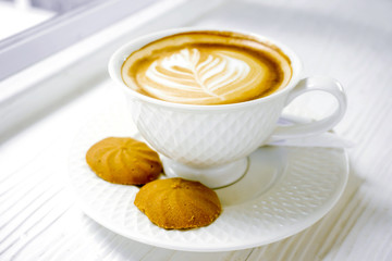 Latte art coffee in a white coffee cup with cookies on white wooden table.