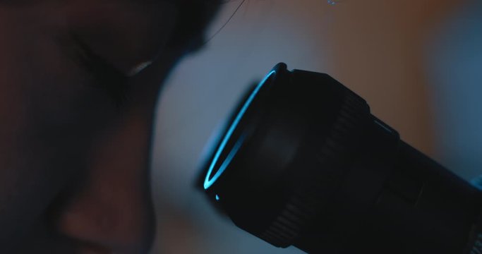 Extreme close up of a female research scientist eyes, looking into microscope.Blue lighting in a dark lab room.Slow motion, slider, shot with BMPCC 4K.Biochemistry, pharmaceutical medicine,science
