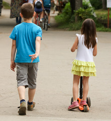 The boy is walking and the girl is riding a scooter