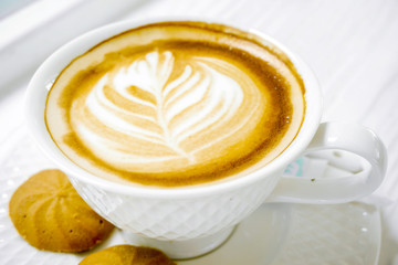 Closeup latte art coffee in a white coffee cup with cookies on white wooden table.