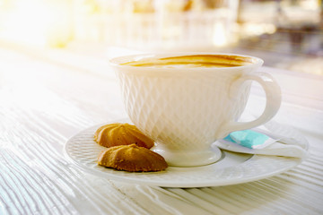 Hot coffee in a white coffee cup with cookies on white wooden table on sun flare background.