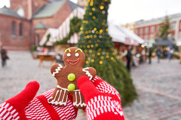 Hand in red mitten holding a smiling gingerbread man and christmas mood in blurred background. Christmas market in old town European small city.
