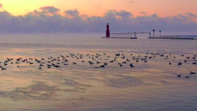Iconic Lighthouse silhouetted against cold, fiery Lake Michigan twilight horizon, flock of geese swimming in frigid waters. Aerial view.