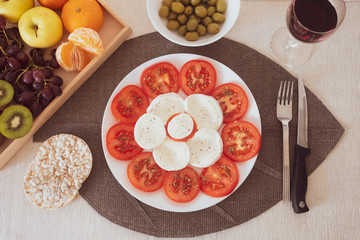 Stay fit with a light and healthy dish of caprese salad with tomatoes and mozzarella. Accompanied by rice cakes, green olives and fruit. A glass of red wine