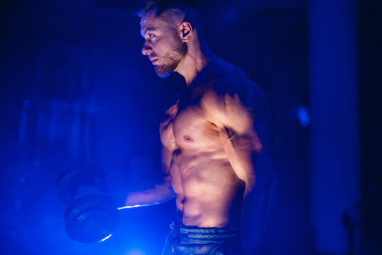 Muscular Young Man Lifting Weights On Dark Background With Blue Light Filter, Naked Torso. Closeup.