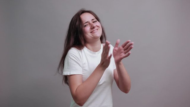 Attractive brunette young woman in fashion white shirt claps hands, showing thumbs at camera isolated on gray background in studio. People sincere emotions, lifestyle concept.