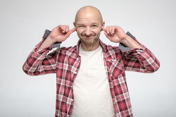 Bald, unshaven man covered ears with index fingers so as not to hear, smiles slyly and looks at the...