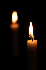 Two isolated burning yellow candles on a black background. The flame of two orange wax candles in the dark