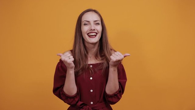 Charming caucasian young woman in maroon dress isolated on orange background in studio claps her hands, pointing thumbs aside. People sincere emotions, lifestyle concept.