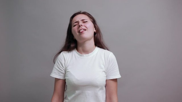 Disappointed caucasian young woman in fashion white shirt showing tongue isolated on gray background in studio. People sincere emotions, lifestyle concept.
