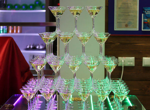 Champagne tower with four rows of glasses. Celebration event party. Bar decoration with led strips of purple and green color lights. Holiday background with shiny drinking glasses. Catering service