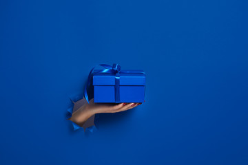 close-up of female hand holding a present gift box through a torn dark blue paper, mockup. Copy...