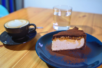 Snickers cheesecake on a blue saucer, a glass of water and a cup of fresh cappuccino on a large bright wooden table