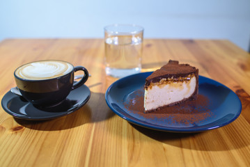 Snickers cheesecake on a blue saucer, a glass of water and a cup of fresh cappuccino on a large bright wooden table