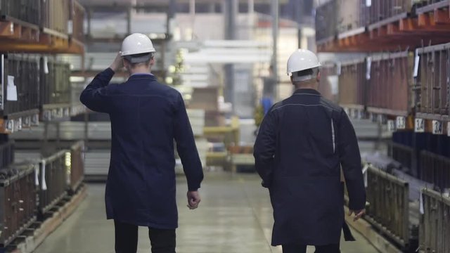 Two Engineers Walking Through Heavy Industry Manufacturing Factory Warehouse. Heavy metallurgical industrial products, manufacturing business production concept. Slow Motion.