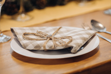 Close-up of ceramic plate with brown and white checked napkin with ribbon on wooden dining table with cutlery. Christmas dinner concept.