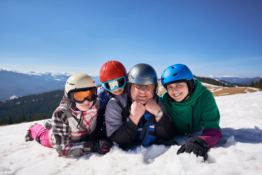 Smiling sportive family, middle-aged man, young woman and kids lying in snow on mountain summit on background of winter mountains and blue sky. Sport, traveling and happy family relations concept. - Powered by Adobe