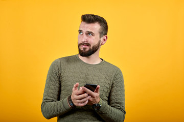 Handsome caucasian man over isolated orange background looking aside, keeping phone in hands, thinking wearing casual green clothes