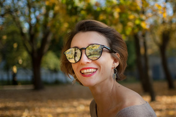 Young and beautiful girl with short hair and sunglasses with mirror surface is posing against the trees with yellow leaves background spending time in the autumn park