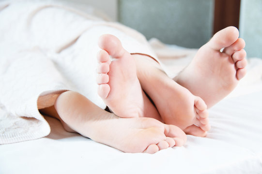 Close-up of the feet of a young couple sticking out from under the covers in the bedroom. Bare feet caress each other engaged in grooming.