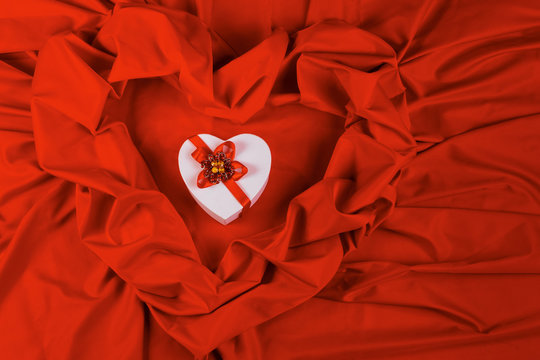 valentines greeting card. white cardboard box in shape of heart wrapped in ribbon lay on a red cloth which repeats the form of present package. love and romance gift concept