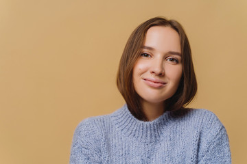 Fototapeta premium Beautiful brunette European woman with makeup, tender smile, dressed in knitwear, enjoys comfort, poses against brown background, copy space area. Lady feels comfort in new winter warm sweater