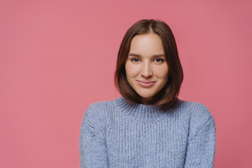 Fototapeta premium Beautiful brunette lady in knitted warm sweater looks directly at camera, smiles positively, enjoys nice winter day, isolated over pink background, has pleasant conversation with interlocutor