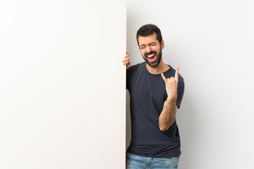 Young handsome man with beard holding a big empty placard making rock gesture