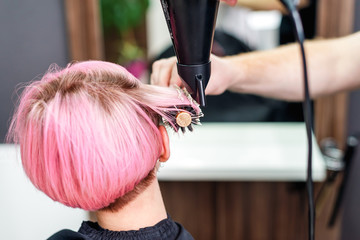 Professional hairdresser drying short pink hair with hair dryer and round brush, close-up.