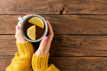 Woman’s hands in knitted sweater holding mug of tea with lemon on wooden background with...