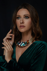 Isolated rich aristocratic makeup woman with brown curls hair in green neckline dress with jewelry necklace holds hands up and touch together and sensuality looks to side with wait on black background