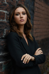 Young brown hair styling fashion woman with beautiful face in black classic coat with opened neck standing in pose with crossed on chest hands arms on the city street with red brick fabric loft wall