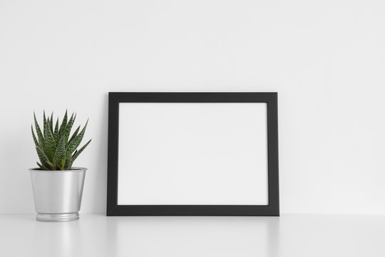 Black frame mockup with a cactus in a pot on a white table.Landscape orientation.