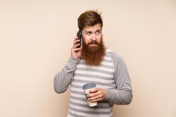 Fototapeta premium Redhead man with long beard over isolated background holding coffee to take away and a mobile