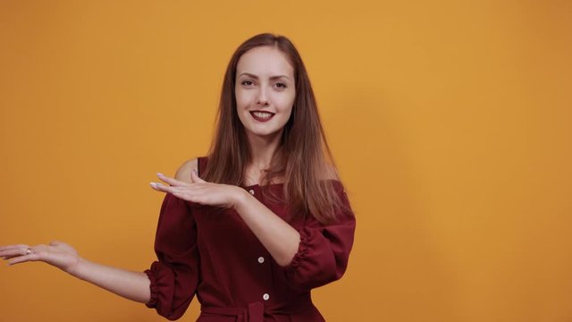 Cheerful charming lady in fashion maroon dress showing space between hands, looking at camera isolated on orange background in studio. People sincere emotions, lifestyle concept.