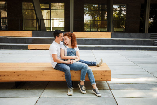 A young couple is sitting on a bench in a modern city, hugging and smiling.