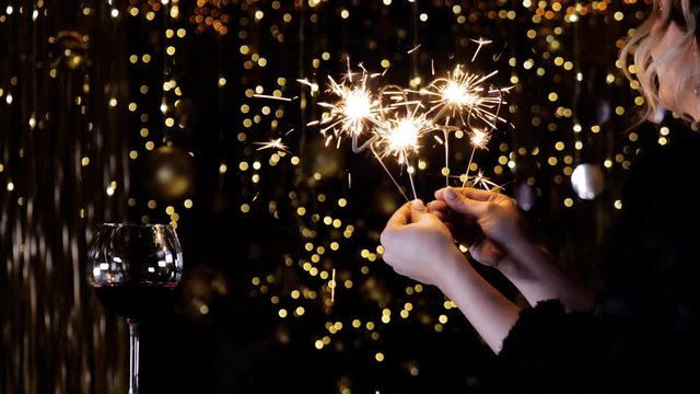 The girl smiles and holds a sparkler in the form of numbers 2020 and sparks fly out. Beautiful shine bengali New Year fire and glass of wine sparkles gold glare. Happy girl waiting fot the New Year.