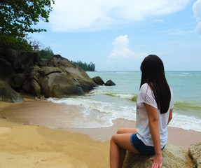 Young attractive tanned girl sitting on a large stone on the beach, admiring the sea scenery