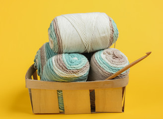 Skein of woolen threads of pastel colors and crochet hook in basket on yellow background....