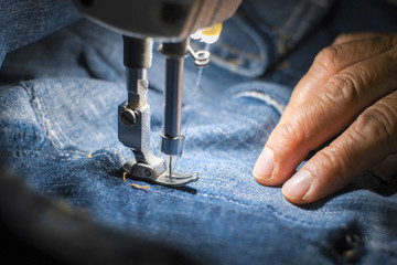 Hand of the seamstress is using a white industrial sewing machine to sew the seams of blue jeans.