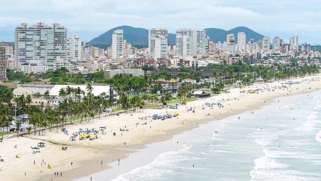 Aerial time lapse of a coastal city on a sunny day at the beach, people on sand and the sea waves hitting the shore. Photo taken at Guaruja SP, Brazil of Enseada beach.