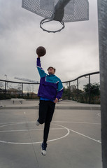 Fototapeta na wymiar Young man dunking/jumping in a outdoor basketball court. Playing basketball. Young man with blonde hair, blue/liliac hoodie and black trousers.