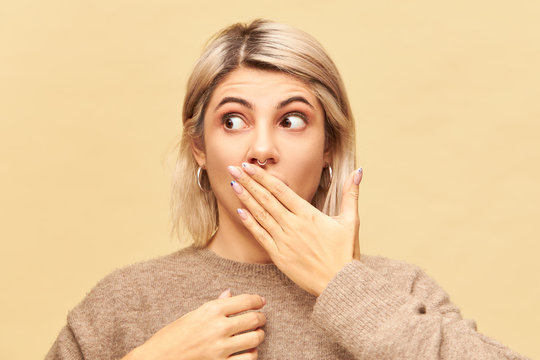 Genuine human emotions, feelings and reaction. Attractive blonde young woman having surprised look, trying to keep secret or confidential information, covering mouth with hand and looking away