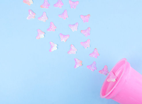 Beautiful pink artificial butterflies flying out of a pink plastic glass (on a blue paper background), minimal concept, flat lay with copy space on the left