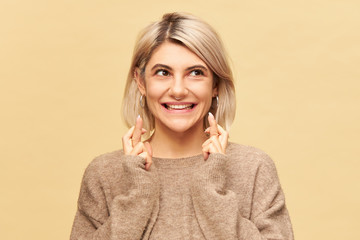 Studio image of attractive stylish student girl in cozy cashmere sweater keeping fingers crossed,...