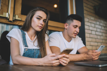Portrait of attractive millennial woman in casual wear looking at camera while spending time with generation boyfriend sitting near and ignoring live communication, concept of cellphone addiction